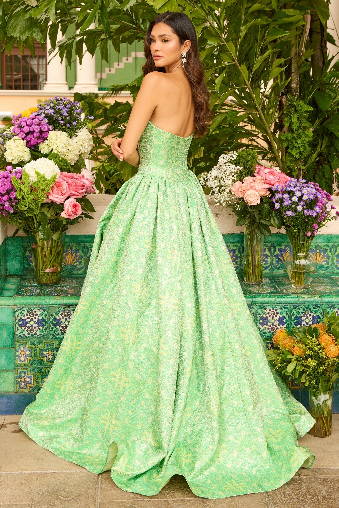 A woman wearing the Ava Presley 42318 long prom dress with a strapless corset bodice stands outdoors among lush greenery and vibrant flower arrangements.